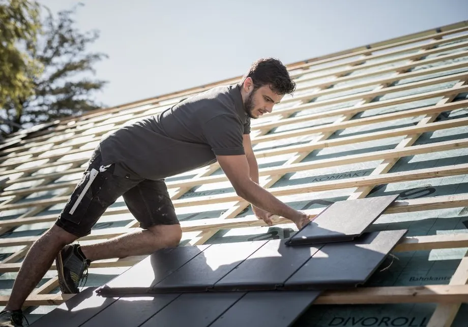 Jumiķis uz jumta ieklāj dakstiņu, fonā redzama jumta konstrukcija ar zemsegumu